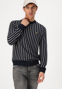 Navy- und weiß gestreifter Pullover mit gerippten Bündchen und Saum. Verfügt über ein kleines gelbes Logo auf der Brust. Getragen mit grauen Jeans.