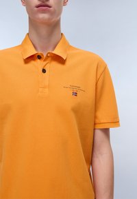 Oranges Polo-Shirt aus strukturiertem Stoff. Mit Kragen, Knopfleiste mit zwei Knöpfen und gesticktem Logo mit norwegischer Flagge.