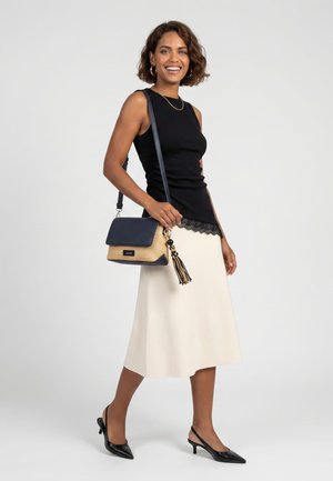 Kvinde smiler, iført sort ærmeløs top med blondekant, hvid midi-nederdel, sorte slingback hæle og bærer en navy og tan crossbody taske.