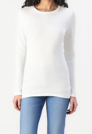 Femme portant un t-shirt blanc uni à manches longues ajusté et un jean bleu clair, debout devant un fond neutre.