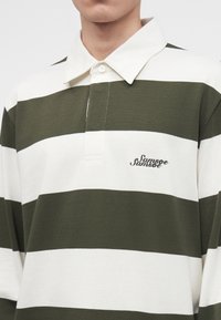 Personne portant un maillot de rugby rayé large vert et blanc avec col blanc et logo "Samsøe Samsøe" sur la poitrine.