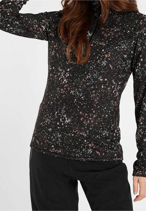 Vrouw die een zwarte top met lange mouwen draagt met een meerkleurig verfspatpatroon en zwarte broek, arm omhoog naar hoofd.