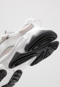 Zapatilla deportiva blanca con elementos de malla y ante, con una suela de goma negra texturizada, conocida por su diseño curvado único y acentos.