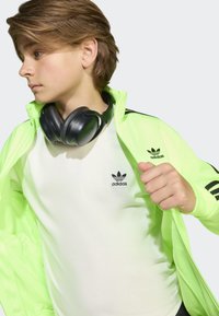 Neongrön sportjacka med svarta detaljer över en vit skjorta med en liten svart logotyp, och svarta hörlurar runt halsen.