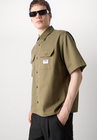 Khaki short-sleeve button-up shirt made of smooth fabric, featuring two chest pockets and a branded tag, paired with black trousers and sunglasses.