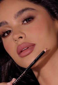 Schwarzer Lidschattenstift mit pinker Spitze, gehalten nahe an vollen, matten Lippen. Natürlicher Hautton mit dezentem Lidschatten und definierten Augenbrauen.