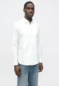 Fehér hosszú ujjú ing gombos gallérral, finom függőleges csíkokkal és sima textúrával, világoskék farmernadrággal párosítva.