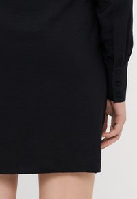 Melna kleita, kas izgatavota no teksturēta auduma, ar garām piedurknēm un pogājamiem aprocēm, taisnu siluetu un līdz ceļgaliem garu malu.