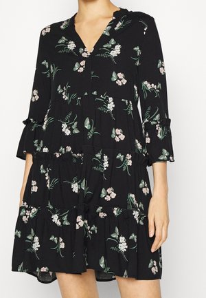 Sort lagdelt kjole med trekvartærmer med grønne og hvide floral- og sommerfugleprint, båret af en stående person.