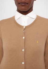 Beige pletený cardigan s kulatým výstřihem, obsahující pět bílých knoflíků a malé žluté logo na levé straně. Pod ním je bílá košilová blůza.