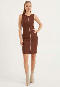 Braunes, geripptes ärmelloses Kleid mit einem Frontreißverschluss und silbernem Zubehör. Das Modell trägt beigefarbene Knöchelstiefel. Glatte Textur, enganliegende Form.