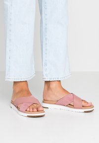 Pink cross-strap sandals with a soft suede texture, a white synthetic sole, and a tan footbed, paired with light blue cropped jeans.