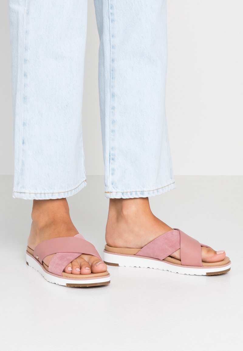 Pink cross-strap sandals with a soft suede texture, a white synthetic sole, and a tan footbed, paired with light blue cropped jeans.