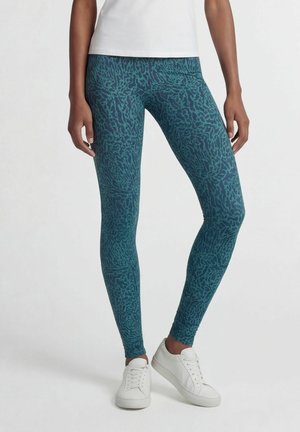 Beine, die blaue gemusterte Leggings und weiße Turnschuhe mit einem weißen Oberteil tragen, stehen vor einem schlichten hellen Hintergrund.
