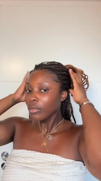 Cheveux tressés en queue de cheval, colliers en or avec des charms, et une serviette blanche texturée. Peau lisse sans maquillage visible.