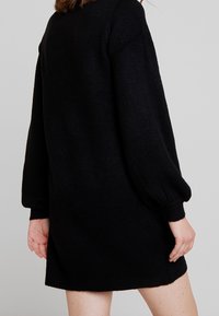 Robe noire tricotée avec des manches longues bouffantes, une coupe ample et des poignets côtelés. Texture lisse avec une apparence douce.