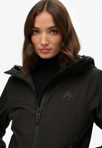 Black waterproof jacket with a zip closure, adjustable hood, and a patch logo on the chest. Smooth, matte texture and fitted design.