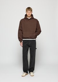Brauner Kapuzenpullover mit Kängurutasche, getragen über einem weißen Shirt, kombiniert mit lockeren schwarzen Jeans und cremefarbenen Sneakers. Einfaches Design.