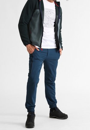 Man in blauwe joggingsbroek, zwarte wandelschoenen, wit T-shirt met grafische afbeelding en groene fleece jas met ritssluiting, met handen in de zakken.