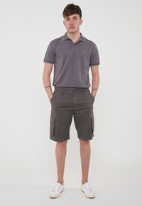 Polo grigio con dettagli turchesi, abbinato a pantaloni cargo grigi con tasche laterali. Indossato con scarpe da ginnastica bianche.