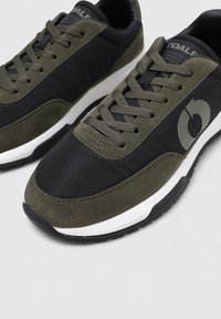 Zapatillas con un upper en negro y verde oliva, que presentan una tela texturizada y acentos de ante. Suela intermedia blanca y suela exterior negra.