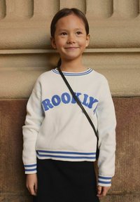Pull blanc avec des accents bleus et le texte "BROOKLYN", doté d'un col rond et de poignets rayés, porté avec une jupe tricotée noire et un sac à épaule.