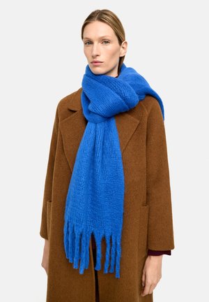 Niebieski szal o teksturowanej powierzchni z frędzlami na końcach, noszony na brązowym płaszczu. Szal ma miękki, dzianinowy wygląd i żywy kolor.