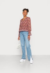 Blusa de padrão floral vermelho com mangas longas, design em forma de v, combinada com jeans azul claro com bainha com fenda; modelo segura uma bolsa branca.