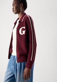 Cardigan a maglia bordeaux con strisce crema, caratterizzato da una grande "G" ricamata sul petto. Dettaglio con tasca con zip e polsini a coste.