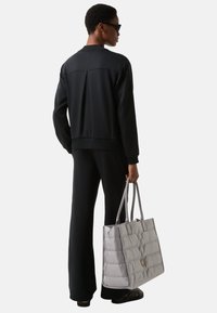 Schwarze, gerippte Strickjacke mit einer Rückenfalte, getragen über passenden Hosen. Trägt eine hellgraue, gesteppte Tragetasche mit zwei Griffen.