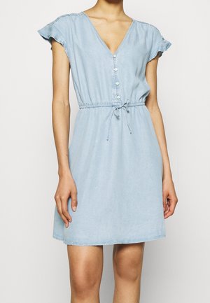Femme portant une robe bleu clair à manches courtes, boutonnée sur le devant, avec une taille à cordon et des détails volantés aux épaules.