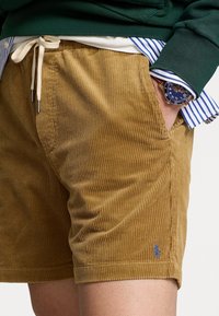 Man som har på sig tanfärgade kordshorts med snörning, grön tröja, blårandig skjorta och armbandsur, med handen i fickan.