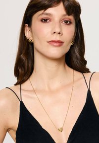 Złota zawieszka w kształcie serca na cienkim łańcuszku, noszona z czarną sukienką z cienkimi ramiączkami. Kolczyki w kształcie serca w kolorze złotym.