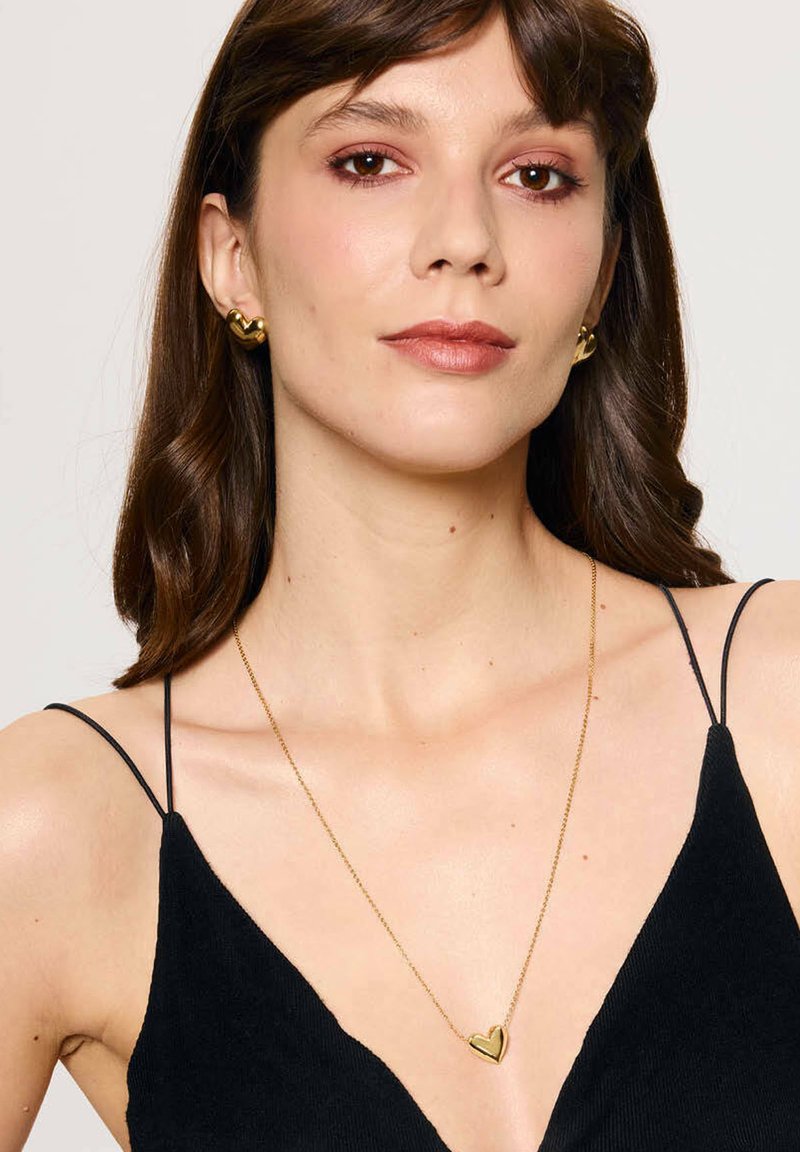 Złota zawieszka w kształcie serca na cienkim łańcuszku, noszona z czarną sukienką z cienkimi ramiączkami. Kolczyki w kształcie serca w kolorze złotym.