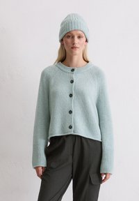 Cardigan corto in maglia azzurro chiaro con polsini a coste, dotato di bottoni neri sul davanti. Abbinato a pantaloni scuri a righe e a un berretto coordinato.