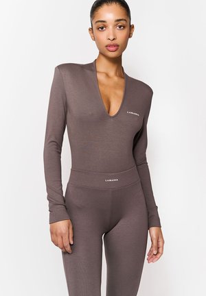 Nő, aki testhez simuló, V-nyakú, hosszú ujjú felsőt és magas derekú leggingset visel tompa barna színben, mindkettő "LAMANIA" márkájú, fehér háttér előtt áll.
