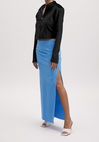 Frau trägt eine schwarze kurze Jacke, einen blauen Maxirock mit hohem Schlitz und weiße Sandalen mit offenem Zeh und Absatz vor einem schlichten Hintergrund.