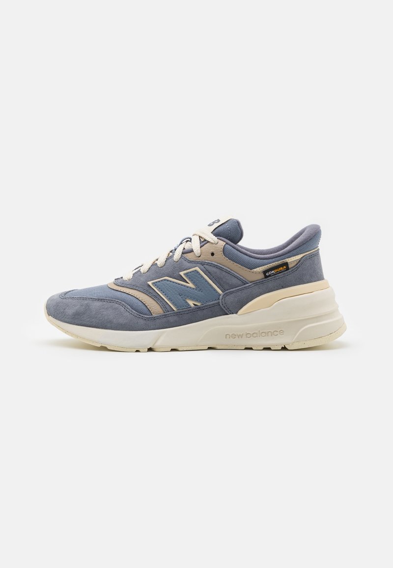 Baskets New Balance bleues et beiges avec tige en daim et mesh, semelle rembourrée, fermeture à lacets, et étiquette en tissu Cordura sur le côté.