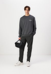 Felpa grigia con polsini a costine e girocollo, abbinata a pantaloni neri a righe sottili con texture e sneaker bianche. Borsa nera tenuta in mano.