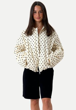 Jeune femme aux longs cheveux bruns porte une veste blanche zippée à pois noirs et un short noir jusqu'aux genoux, debout face à l'avant.