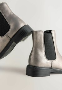 Metallisch zilveren enkelboots met zwarte elastische zijpanelen, een slanke zwarte rubberen zool en contrasterende stiksels. Ontwerp met ronde neus.