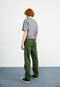 Grijze t-shirt met korte mouwen, gecombineerd met loszittende groene cargobroek met grote zakken, gedragen met donkere sneakers, tegen een eenvoudige achtergrond.