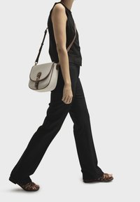 Crossbody taske i lysegrå stof med brune læderdetaljer, prydet med en klaplukning og hardware i guldfarve, båret af en person.