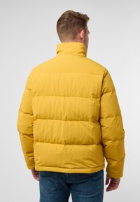 Geel pufferjack met horizontale quiltstiksels, hoge kraag en een gladde textuur. Zichtbare naden en een ontspannen pasvorm. Draagt over blauwe jeans.