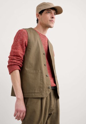 Olive green vest with two front pockets, worn over a textured red sweater. Beige cap and button details enhance the layered look.