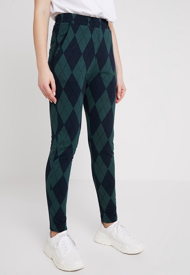 Personne portant un pantalon à motif argyle vert et bleu marine avec une taille élastique, associé à des baskets blanches épaisses et un haut blanc.