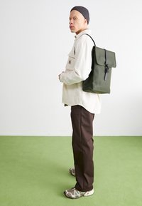 Sac à dos vert à rabat avec quincaillerie noire, forme rectangulaire et finition texturée, porté par une personne en chemise blanche et pantalon marron.