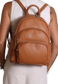 Zaino in pelle marrone con superficie strutturata, parte superiore arrotondata e due tasche frontali con zip. Spallacci regolabili e una maniglia superiore per il trasporto.