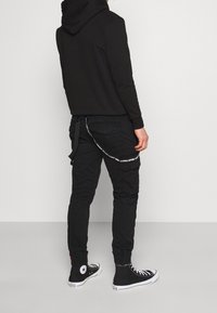 Svart hoodie med en framficka, tillsammans med svarta cargo-byxor med en sidficka och ett vitt band som står "REMOVE BEFORE FLIGHT". Svarta högskor.