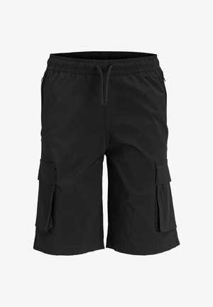 Ragazzo adolescente in piedi, indossa una felpa con cappuccio giallo pallido, pantaloncini cargo neri, calzini bianchi con marchio e scarpe da ginnastica bianche, guarda verso destra e sorride.